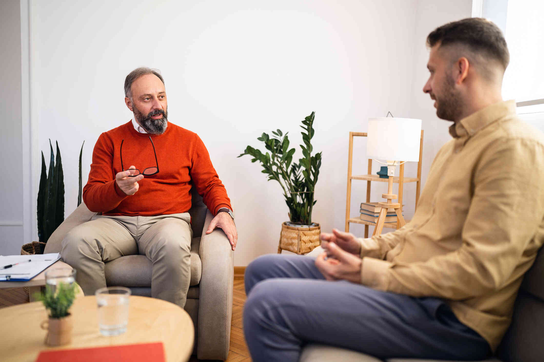 A man in a red shirt holds his glasses as he sits in a chair and speaks to another man sitting across from him in an office.